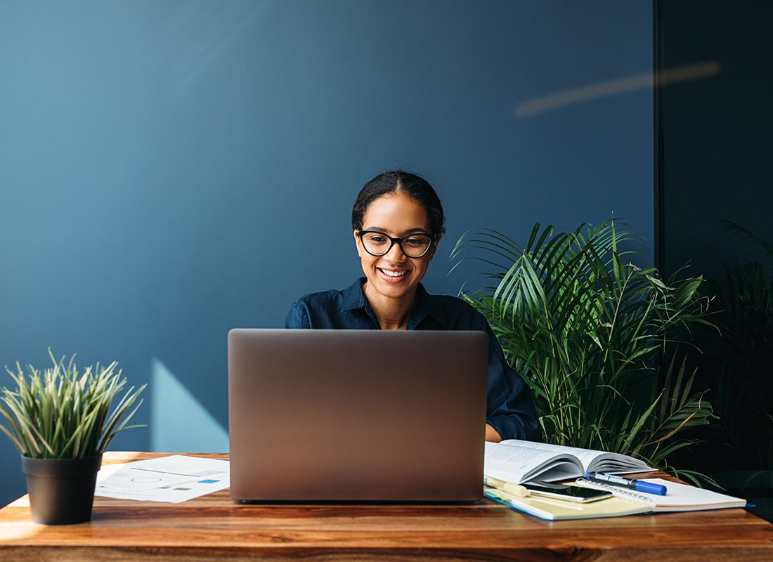 Professional Liability Quote - Professional Woman Smiling and Staring at Her Laptop While She Sits in a Modern Office at Her Desk with Books and Supplies